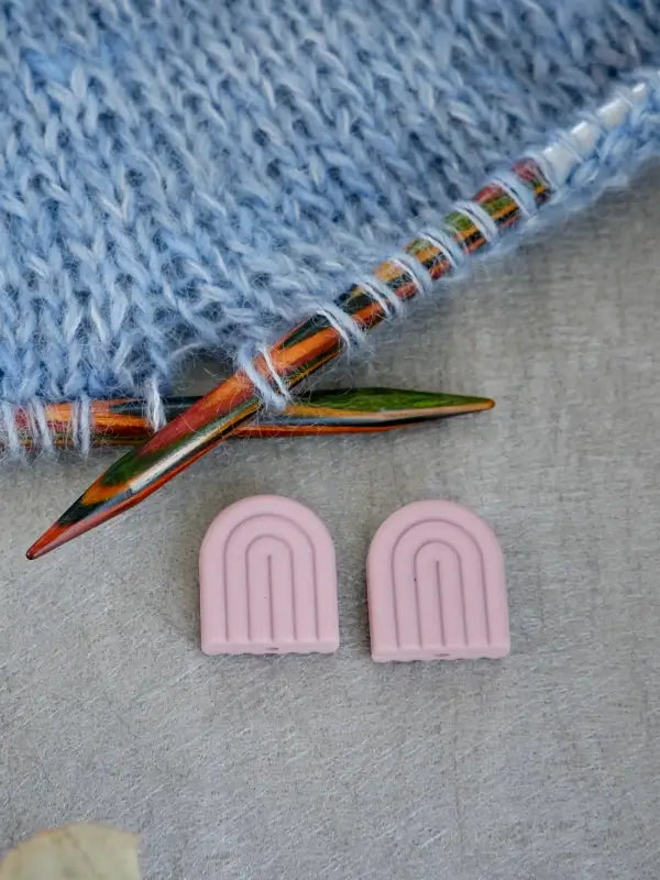 Two colorful wooden knitting needles rest on blue knitted fabric. Below them, two small Woolwomen Puikkostopparit—pink, arch-shaped accessories with grooved patterns—are placed on a neutral surface.