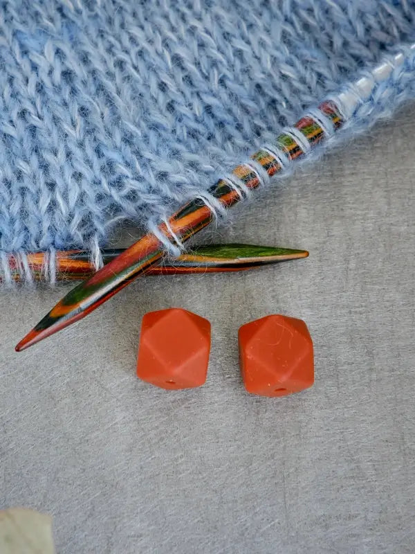 A close-up of two orange Woolwomen Puikkostopparit next to colorful wooden knitting needles and light blue knitted fabric on a gray surface.