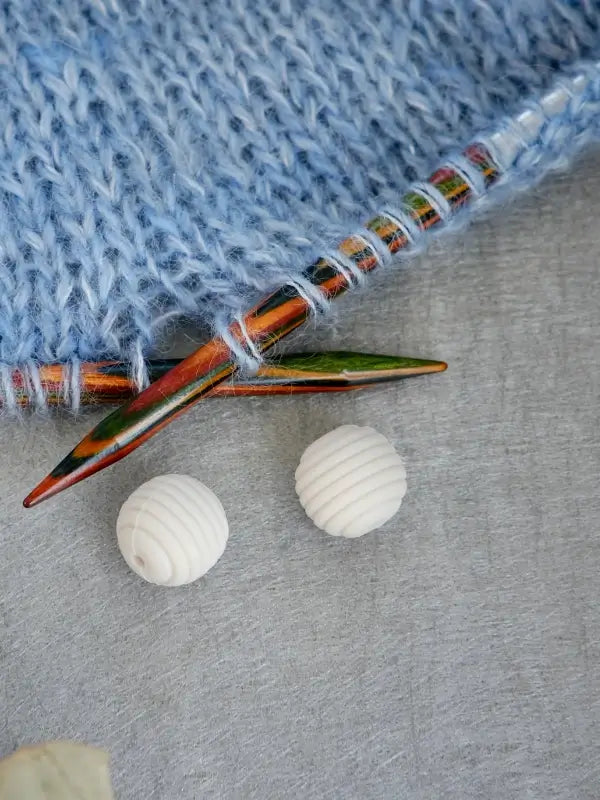 Close-up of colorful knitting needles in light blue yarn, with two white, round Woolwomen Puikkostopparit lying nearby on a gray surface.