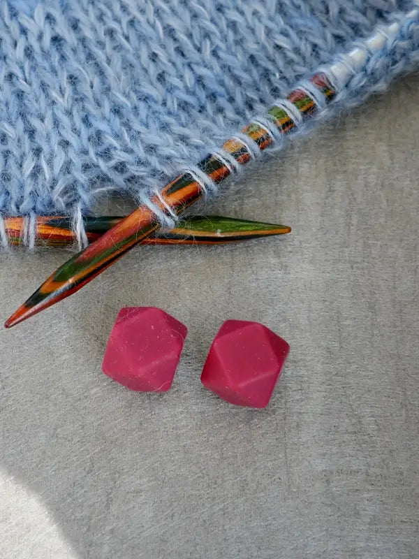 A close-up of two faceted pink dice on a gray surface, next to a blue knitted fabric adorned with Woolwomen Puikkostopparit multicolored knitting needles.