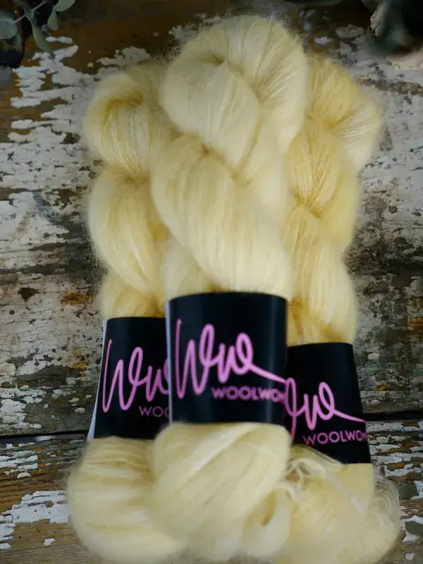 Three skeins of WoolWomen Silk Mohair in pale yellow, each with a black label featuring pink cursive writing, arranged on a rustic wooden surface.