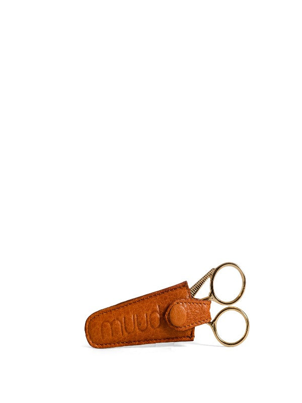 A small pair of gold-colored scissors partially inside a muud Espoo saksikotelo whisky with a button flap, set against a plain white background.