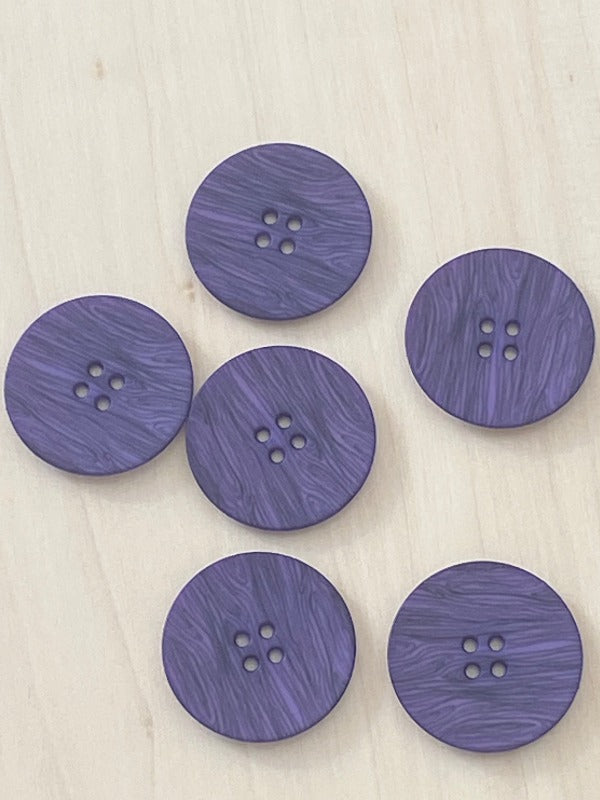 Six round, purple Muovinappi 23mm buttons with a marbled pattern and four holes each are arranged on a light wooden surface; nämä napit ovat kevyet ja saatavilla eri väreissä.