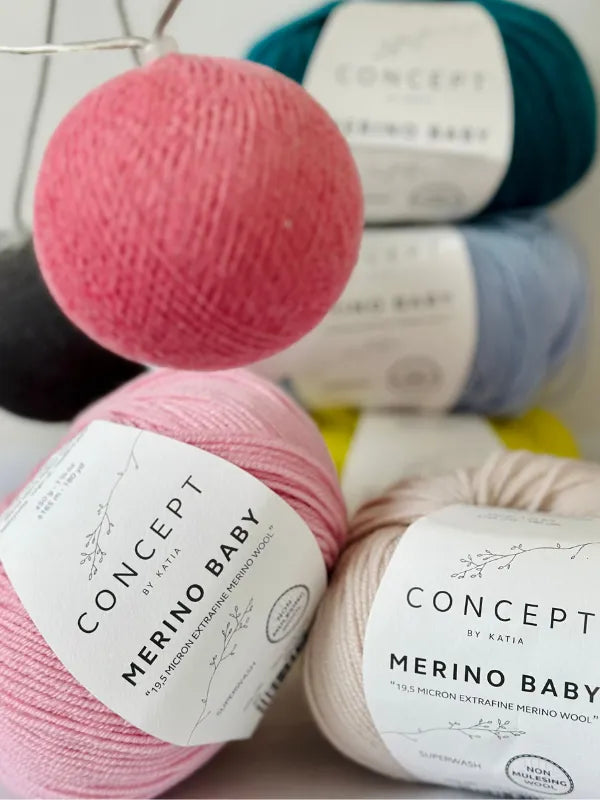 Four balls of Concept by Katia Merino Baby yarn in cream, pink, blue, and teal are arranged together, with a close-up focus on the labels and soft texture of the wool.