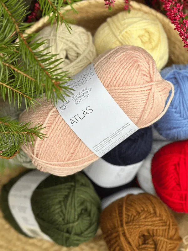 A basket filled with colorful balls of Sandnes Garn Atlas yarn—pink (with an Atlas label), blue, red, and more—sits with a pine branch in the foreground.