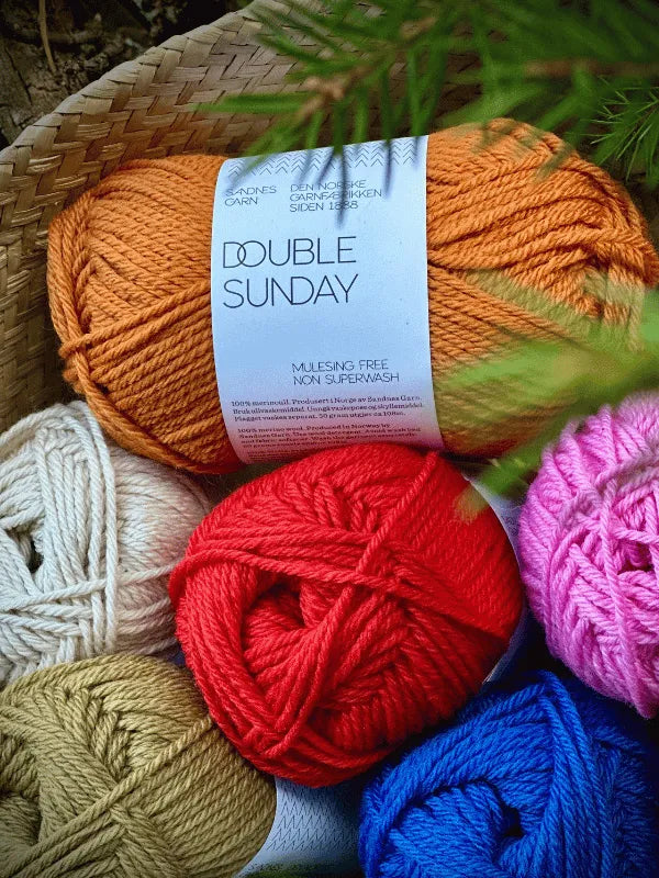 A close-up of colorful skeins of Sandnes Garn Double Sunday yarn, in shades of orange, red, pink, blue, cream, and tan, nestled in a woven basket with green leaves partially covering the edge.