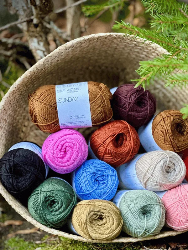 A woven basket filled with colorful Sandnes Garn Sunday fingering-weight yarn in brown, pink, blue, green, black, cream, and orange sits outdoors among greenery and tree branches.