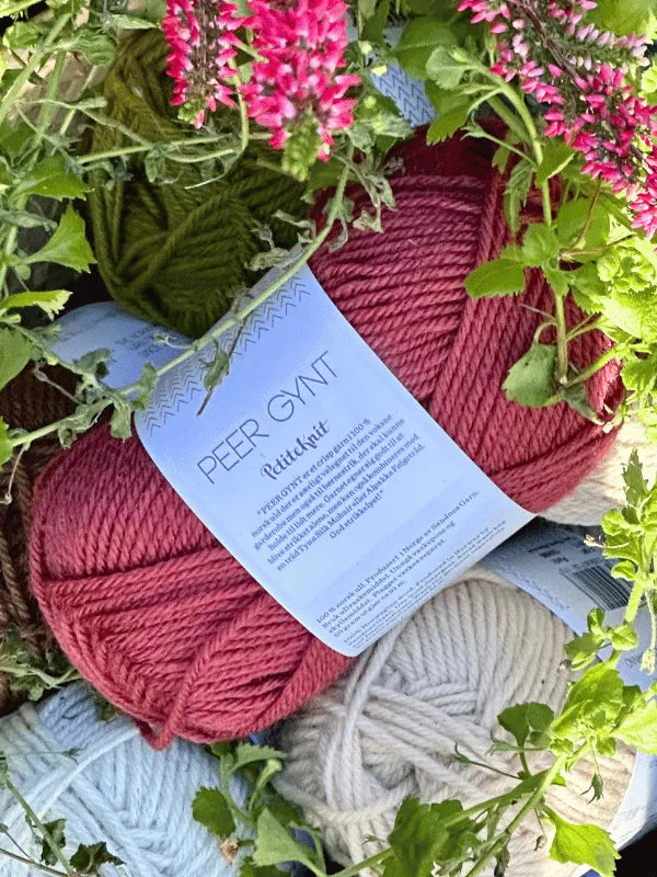 Close-up of Sandnes Garn Peer Gynt skeins in red, white, and green, partially covered by green leaves and pink flowers—ideal for Norwegian wool projects or vibrant colorwork knitting.