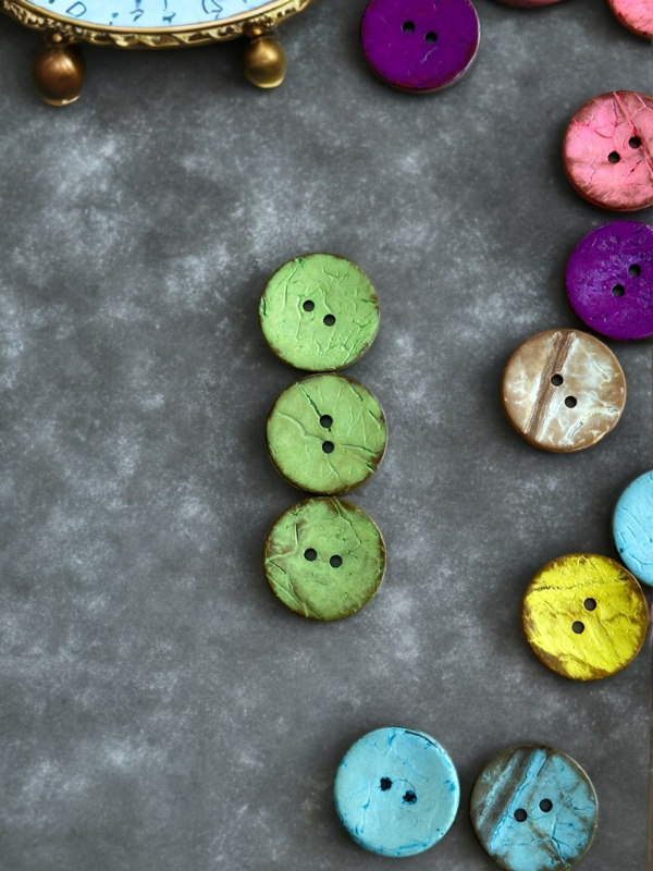 Colorful round buttons, featuring the Kookosnappi värikäs 22m (8871), are displayed on a gray textured background, with three green buttons in a central vertical line and part of a gold-trimmed clock in the top left corner.