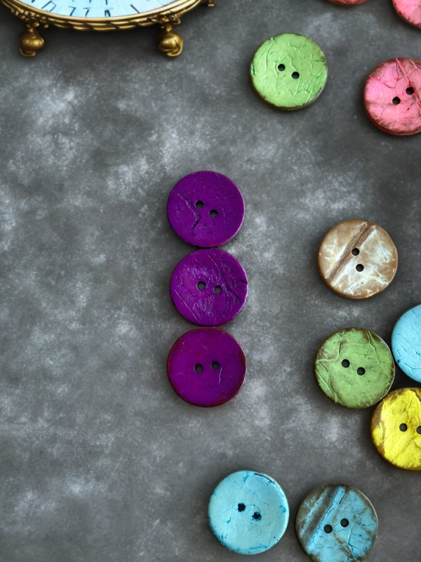 Colorful round buttons, such as the Kookosnappi värikäs 22m (8871), are arranged on a gray surface with four purple buttons in a vertical line at the center. A vintage clock is partly visible in the top left, and more buttons are scattered around.