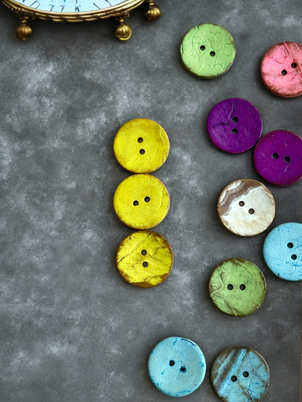 Colorful round buttons, including the Kookosnappi värikäs 22m (8871), are neatly arranged on a gray surface, with part of a decorative clock visible in the top left corner.