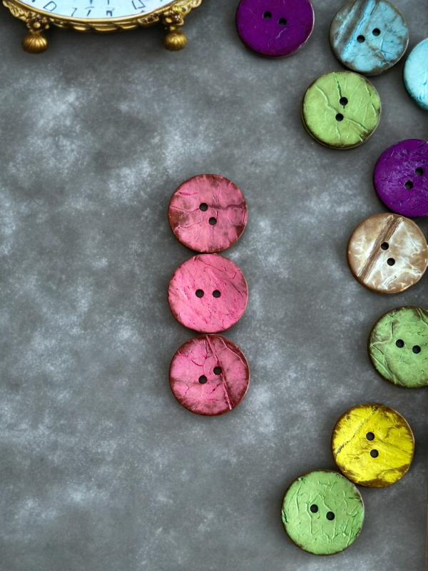 Large, round, colorful Kookosnappi värikäs 22m (8871) buttons are arranged on a mottled gray surface, with four vertically aligned at the center. An ornate clock appears in the upper left corner.