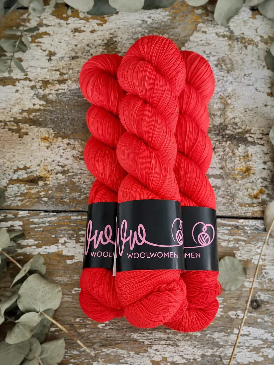 Two skeins of bright red WoolWomen Merino Sock yarn with black labels, resting on a weathered wooden surface.