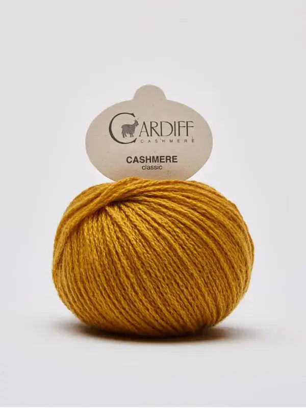 A ball of mustard yellow Cardiff Cashmere Classic – 100 % kashmirlanka rests on a plain surface with its label visible, highlighting the luxurious texture of this premium kashmirlanka yarn.