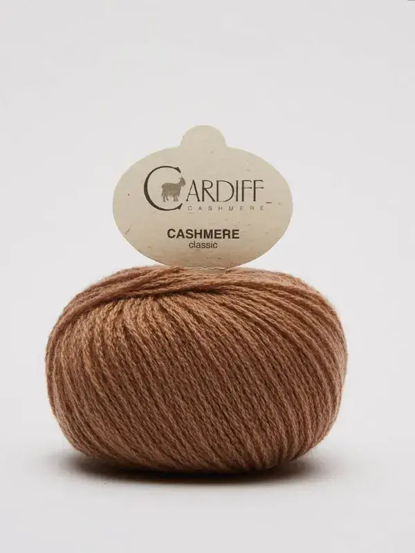 A ball of light brown Cardiff Cashmere Classic – 100 % kashmirlanka rests on a white background, with its product label attached at the top, highlighting the luxurious quality of this premium cashmere yarn.