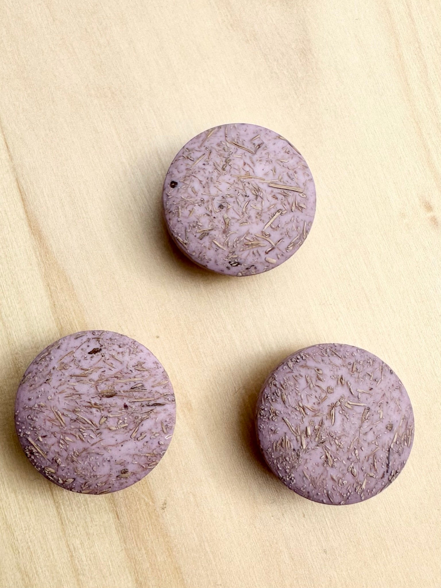 Three round, purple Kierrätysnappi kookos 20mm (0454241) buttons with textured surfaces are arranged in a triangle on a light wooden surface.