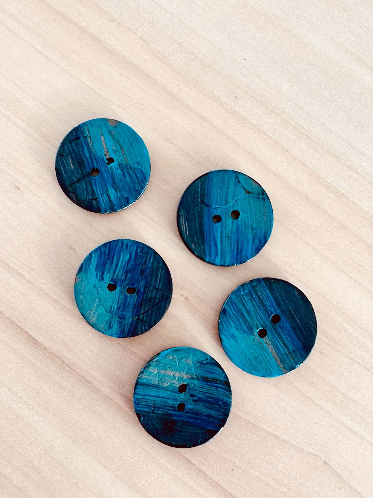 Five round, blue Käsinmaalatut kierrätysnapit 25mm (0454171) with two holes each are arranged loosely on a light wooden surface, showing textured, painted finishes in varying blue shades.