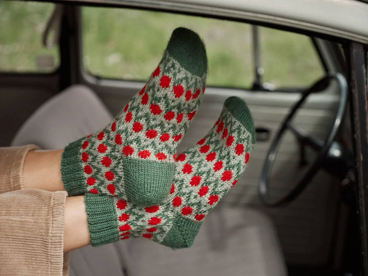 A person’s feet resting on a car dashboard, wearing green wool socks featuring a red and cream geometric pattern from the Jenna Kostet - Neulottu Kalevala III, Ennakkotilaus collection.