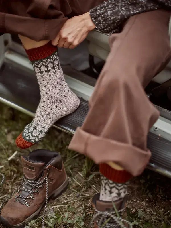 A person putting on Jenna Kostet - Neulottu Kalevala III, Ennakkotilaus patterned knit socks with red cuffs and toes while sitting near brown hiking boots.