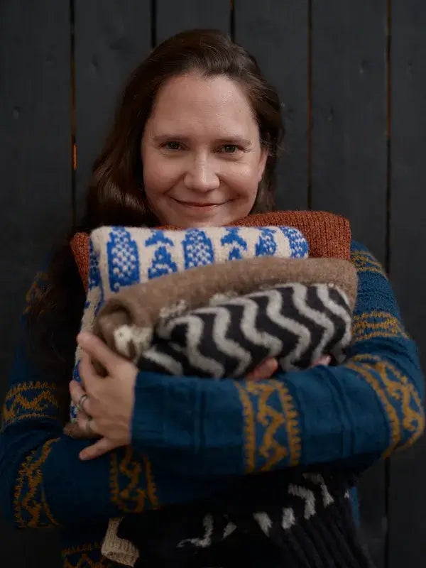 A woman smiling while holding a stack of folded sweaters from the Jenna Kostet - Neulottu Kalevala III, Ennakkotilaus collection.