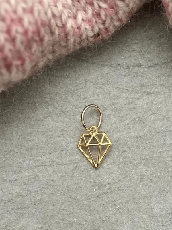A small gold wire charm from KNITS by cindy ekman silmukkamerkit, shaped like a geometric diamond, rests on grey beside neulonta-inspired pink and cream knit fabric—ideal for anyone looking for kauniit silmukkamerkit.
