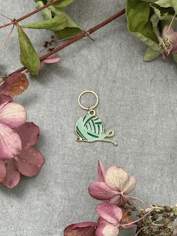 The KNITS by cindy ekman silmukkamerkit is a mint green yarn ball keychain with knitting needles, ideal for neulonta lovers. Shown on a gray surface with pink and green dried flowers and leaves.