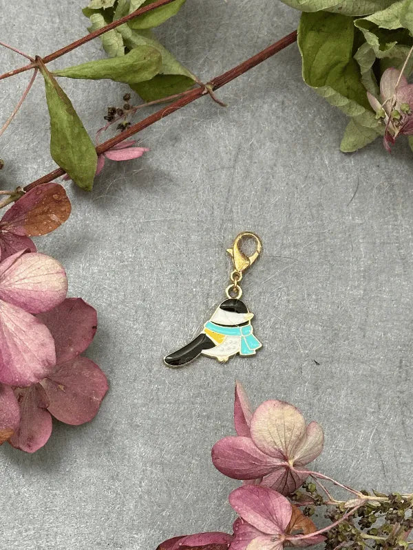 A KNITS by cindy ekman silmukkamerkit charm—shaped like a person in traditional Korean hanbok with a gold clasp—rests on gray amid dried pink and green flowers, ideal as kauniit silmukkamerkit for neulonta projects.