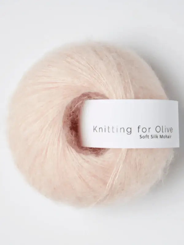 A ball of Knitting for Olive Soft Silk Mohair yarn, in a delicate pale pink shade and spun from kid mohair and silkki lanka, rests on a plain white background. The skein features a white label printed with “Knitting for Olive Soft Silk Mohair,” highlighting its luxurious quality and Responsible Mohair Standard certification.