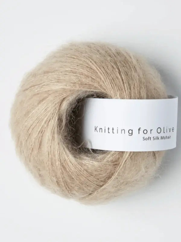 A ball of Knitting for Olive Soft Silk Mohair yarn in beige rests on a light surface, highlighting its soft and fluffy texture. This luxurious silkki lanka is crafted from Responsible Mohair Standard-certified kid mohair, offering both superior softness and ethical sourcing.
