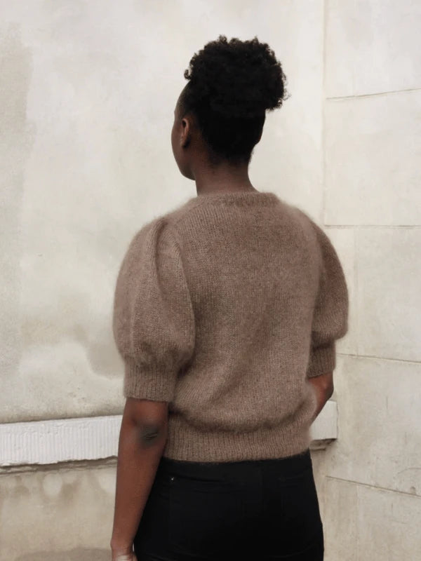 A person with dark skin and curly hair in a puff hairstyle faces away, wearing a brown Puff Tee neuleohje short-sleeved knit sweater and black pants, standing against a light-colored textured wall.