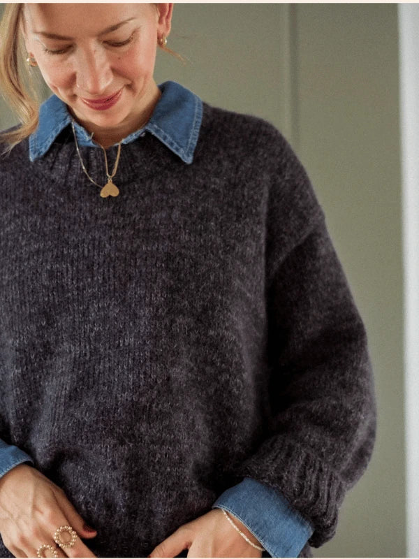 Wearing the KutovaKika Cinema Paita sweater, styled over a blue denim shirt and accessorized with a heart necklace, rings, and a bracelet, this person gazes downward with a gentle smile.