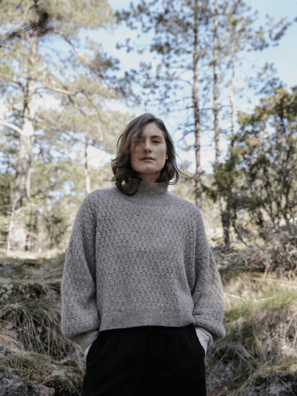 A person with shoulder-length hair wears a gray textured Laine Juhlanumero - ENNAKKO neulekaavat sweater and black pants while standing in a forest, capturing pohjoismainen estetiikka with tall trees and a clear sky in the background.