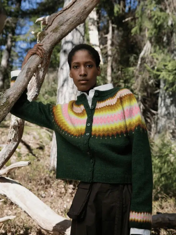 Outdoors in a forest, a person leans on a tree branch wearing the Laine Juhlanumero - ENNAKKO: a green neulekaavat sweater with colorful patterns and a white collared shirt, capturing pohjoismainen estetiikka with a serious expression.