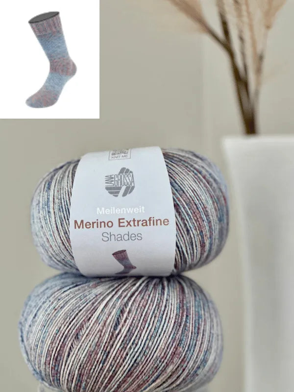 Two skeins of Lana Grossa Meilenweit 100 Merino extrafine Shades, a mulesing-free merino sock yarn in soft pastels, stacked with visible labels. Inset shows a knitted sock from the same gradient-dyed yarn. Blurred background with white vase and dried plant.