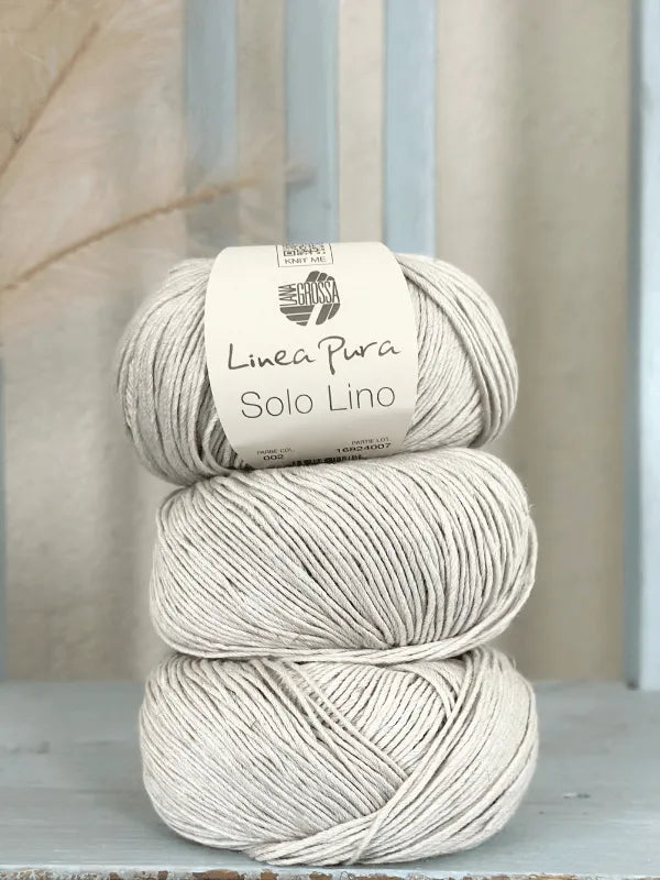Three skeins of beige Lana Grossa Solo Lino, a vegan linen yarn, are stacked against a light blue wooden background. The top skein features a white label with the brand and product information.