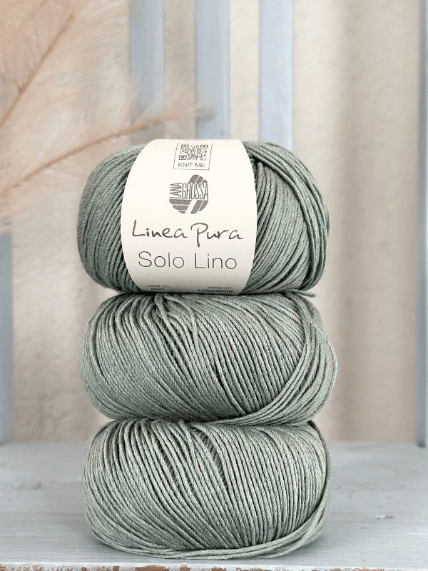 Three sage green balls of yarn are stacked vertically, each with a beige label reading Lana Grossa Solo Lino. A softly blurred neutral background highlights this vegan yarn’s natural beauty.