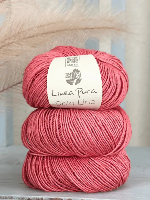 Three skeins of Lana Grossa Solo Lino, each wrapped with a beige label, are stacked on a light surface. Soft beige pampas grass blurs in the background, accentuating this vegan yarn’s delicate beauty.