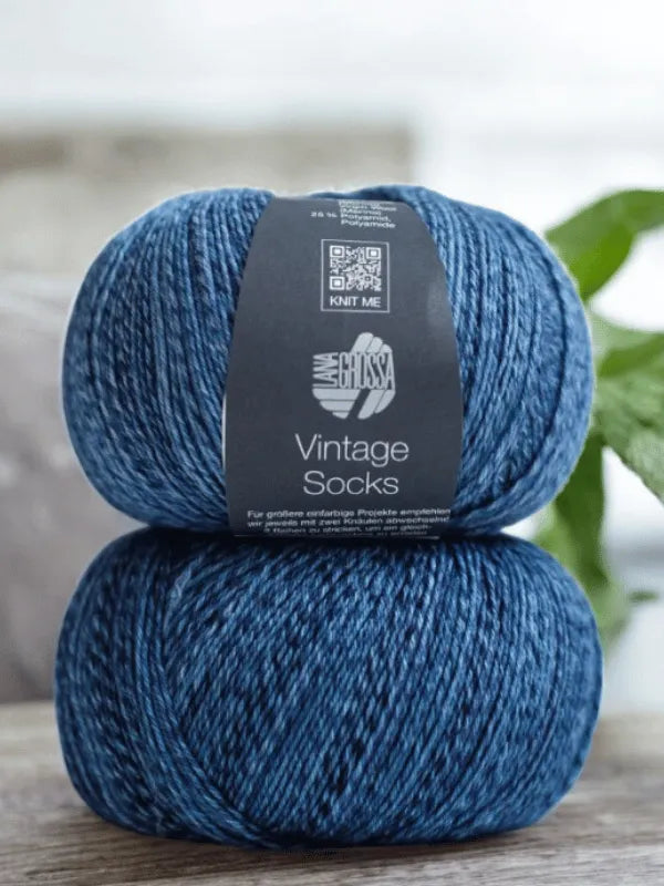 Two skeins of Lana Grossa Vintage Socks yarn from Lauri Fingerin sukkaohje + Lana Grossa Vintage socks are stacked, their labels showing "Vintage Socks" and "Lauri sukkaohje," with a blurred green plant in the background.
