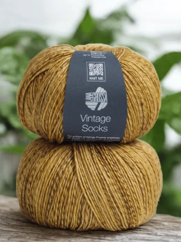 Two mustard-colored Lana Grossa Vintage Socks yarn balls are stacked on wood, perfect for your next Lauri Fingerin sukkaohje + Lana Grossa Vintage socks project, with blurred green plants in the background.