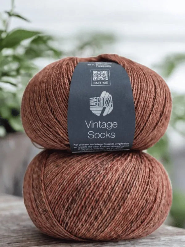Two balls of Lana Grossa Vintage Socks yarn from the Lauri Fingerin sukkaohje +Lana Grossa Vintage socks set sit on wood, ideal for knitting villasukat, with blurred green plants in the background.