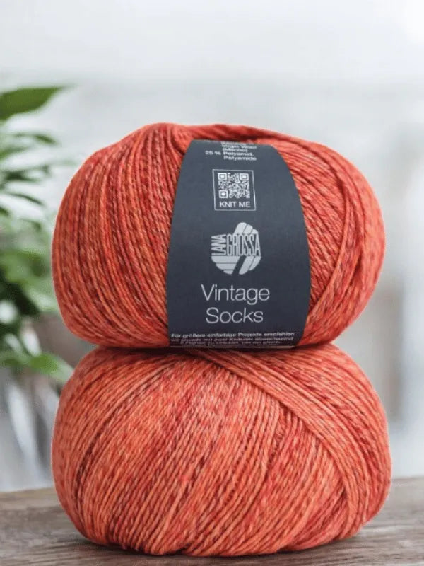 Two balls of Lana Grossa Vintage Socks yarn from the Lauri Fingerin sukkaohje +Lana Grossa Vintage socks set are stacked on wood, ready for your next villasukat or sukkaohje project, with a blurred plant in the background.