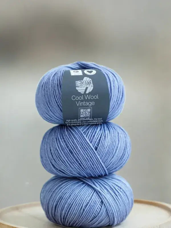 Three skeins of blue Lana Grossa Cool wool vintage yarn stacked vertically, with a product label wrapped around the top skein.