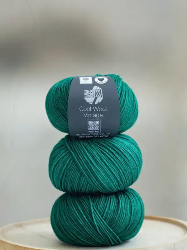Three stacked skeins of dark emerald green Lana Grossa Cool wool vintage yarn with a product label wrapped around the top skein.