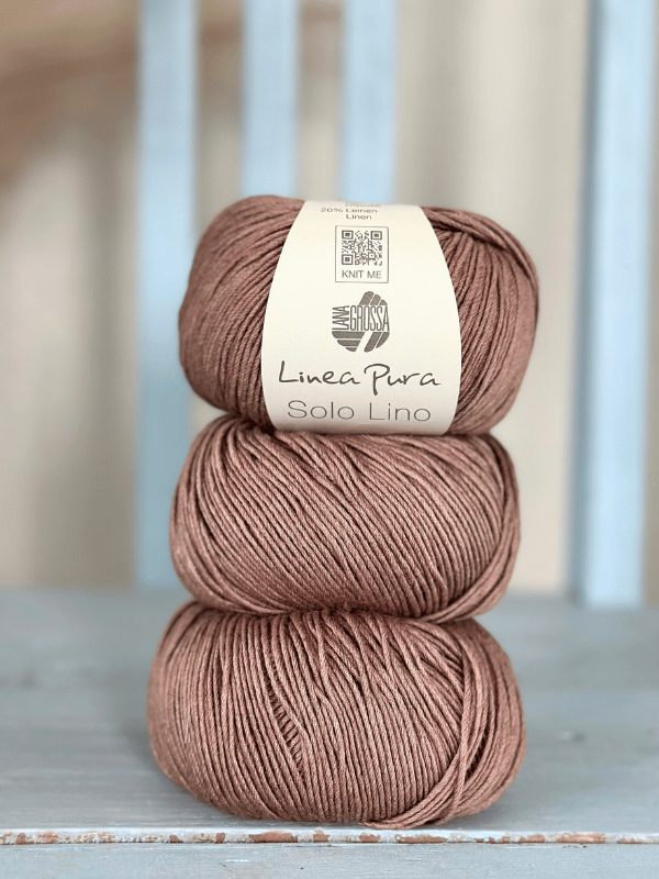 Three balls of light brown Lana Grossa Solo Lino yarn are stacked on a wooden surface. The top ball shows its label. In the background, soft vertical blue slats appear blurred.