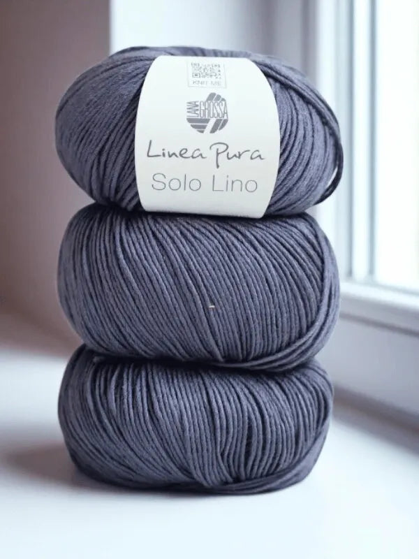 Three skeins of gray Lana Grossa Solo Lino, a vegan yarn, are stacked on a white windowsill with natural light streaming in through the window.