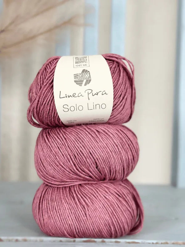 Three skeins of dusty pink Lana Grossa Solo Lino are stacked, the top one wrapped in its label. Softly blurred neutral tones in the background highlight this vegan yarn choice.