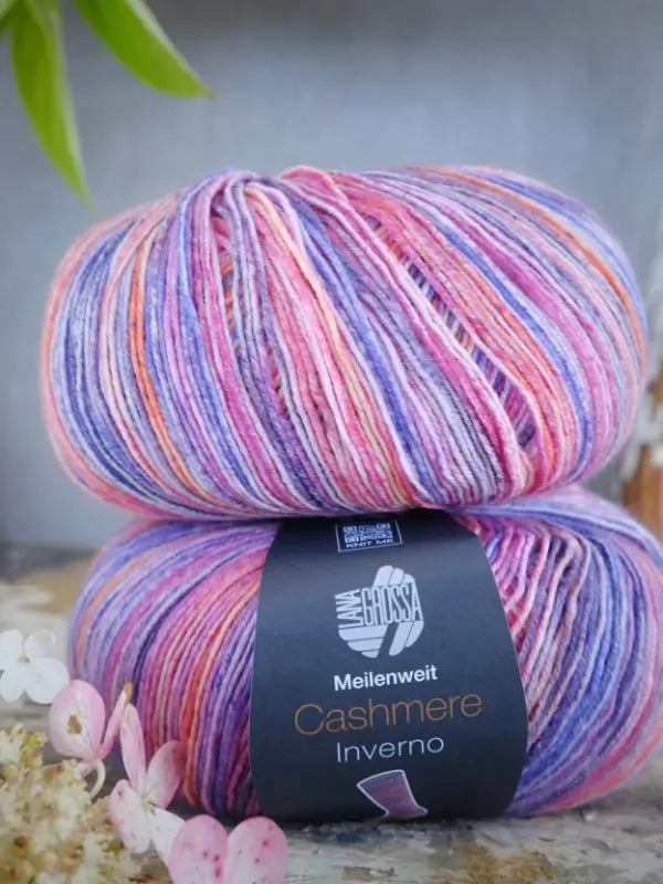 Two skeins of multicolored yarn in pastel shades of purple, pink, orange, and white, labeled Lana Grossa Meilenweit 100 Cashmere Inverno, with flowers in the foreground.