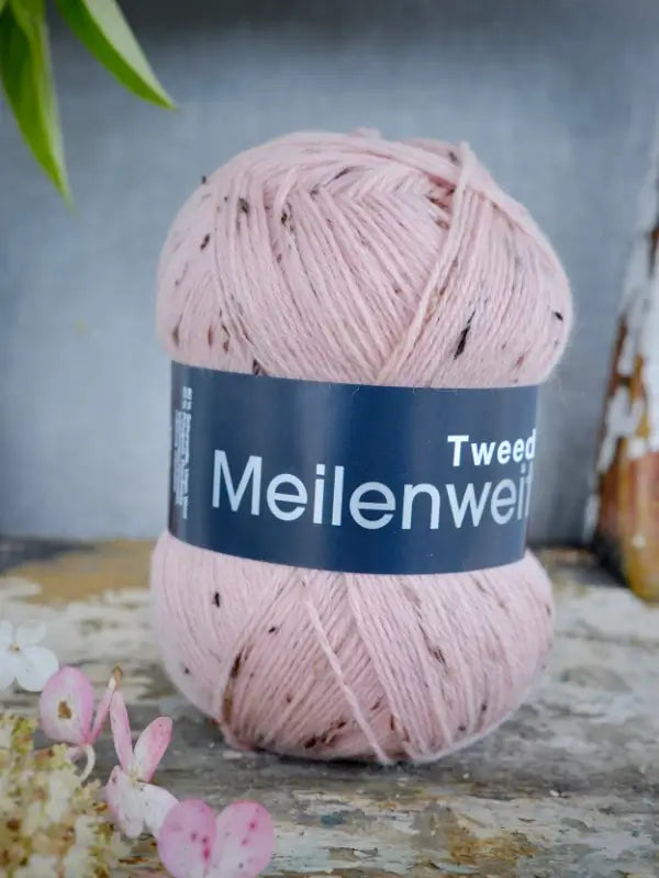 A skein of Lana Grossa Meilenweit 100 Tweed yarn in light pink with subtle dark flecks rests on a rustic surface, accompanied by small flowers—ideal for your next sukkalanka project.