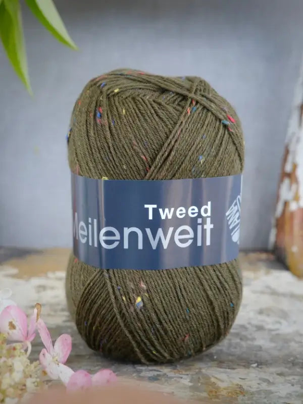 A skein of Lana Grossa Meilenweit 100 Tweed yarn in olive green with colorful flecks rests on a surface. The label displays the product name, and small pink flowers appear in the lower left corner, while a rustic wooden object is visible to the right, accentuating this charming tweed sukkalanka yarn.