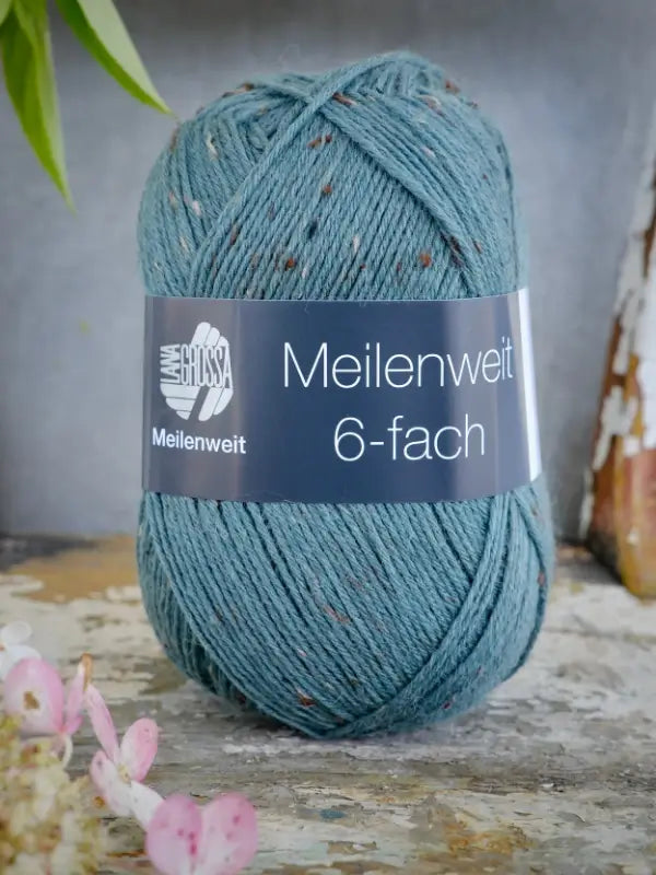A ball of Lana Grossa Meilenweit 6 Tweed yarn in a blue-green speckled color rests on a surface, with flowers in the foreground and green leaves to the left, highlighting the subtle tweed-like texture of the yarn.